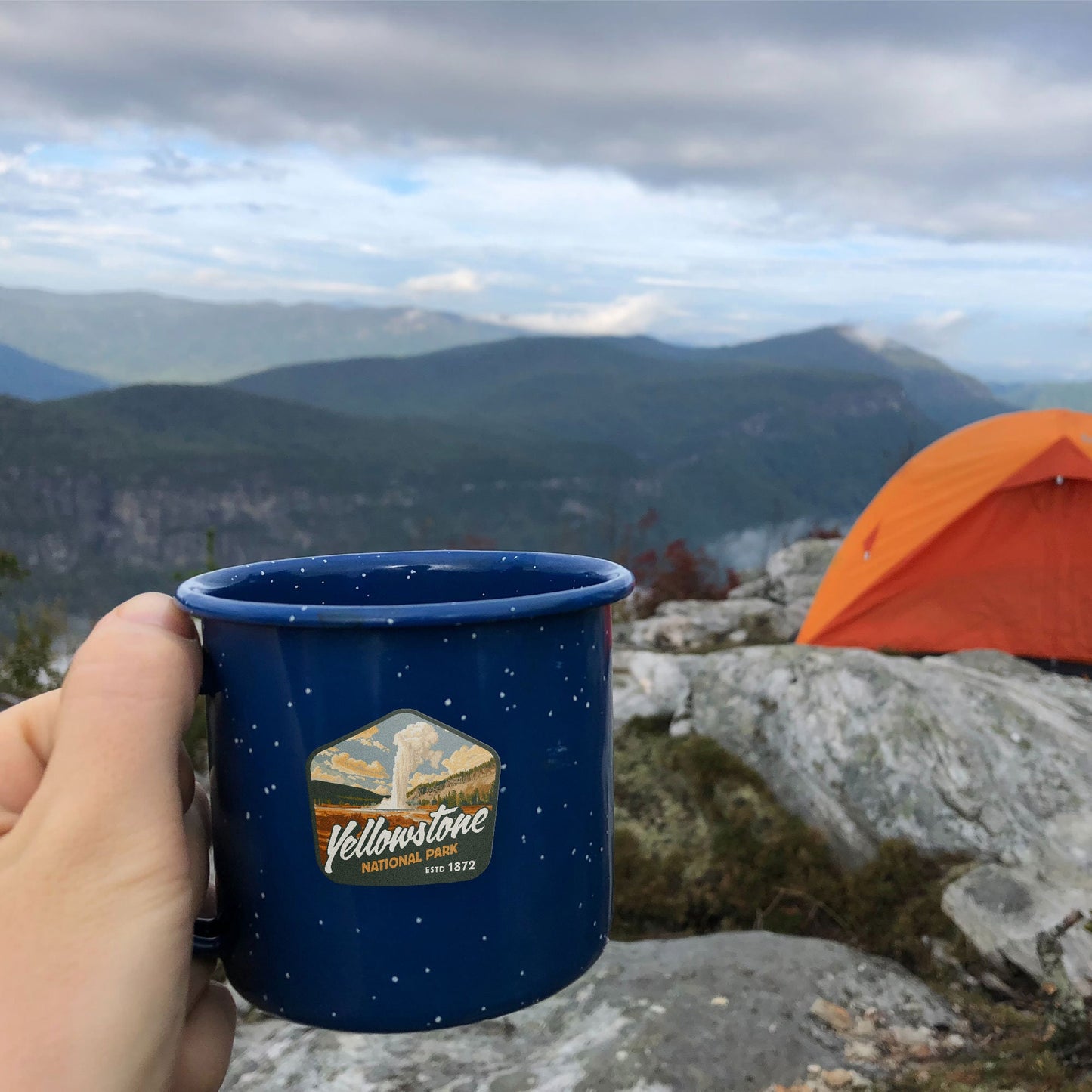 Waterproof vinyl sticker of Yellowstone National Park on blue camping mug with orange REI tent in background. Features captivating mountain art. Perfect hiking sticker for outdoor enthusiasts and adventurous travelers.